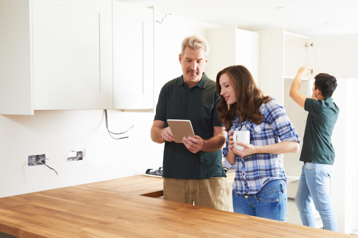 Two people in an unfinished kitchen looking at a tablet while a person installs a cabinet behind them