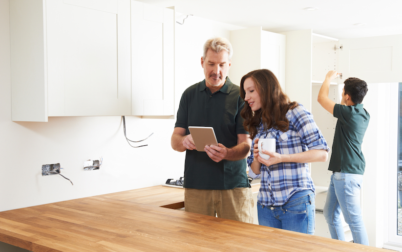 Two people in an unfinished kitchen looking at a tablet while a person installs a cabinet behind them