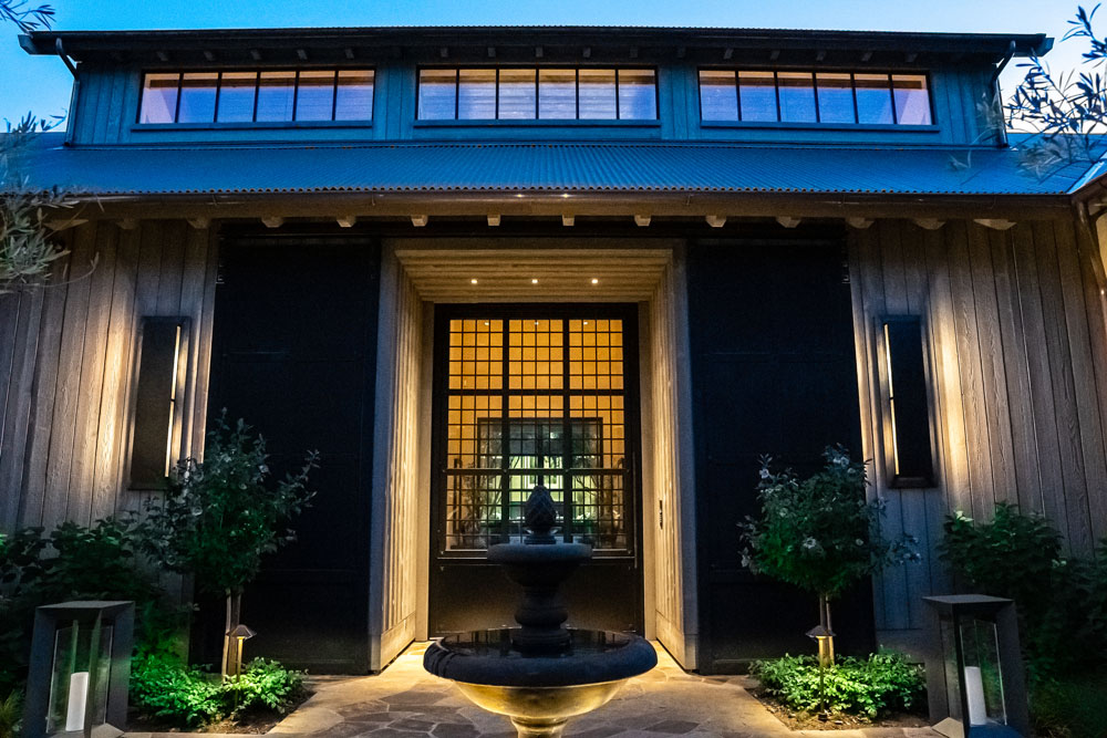 In the middle of this exterior shot is an illuminated bird feeder. Two young trees flank a window. 