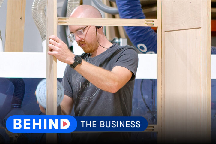 Construction worker building a cabinet in a shop with text that reads "Behind the Business."