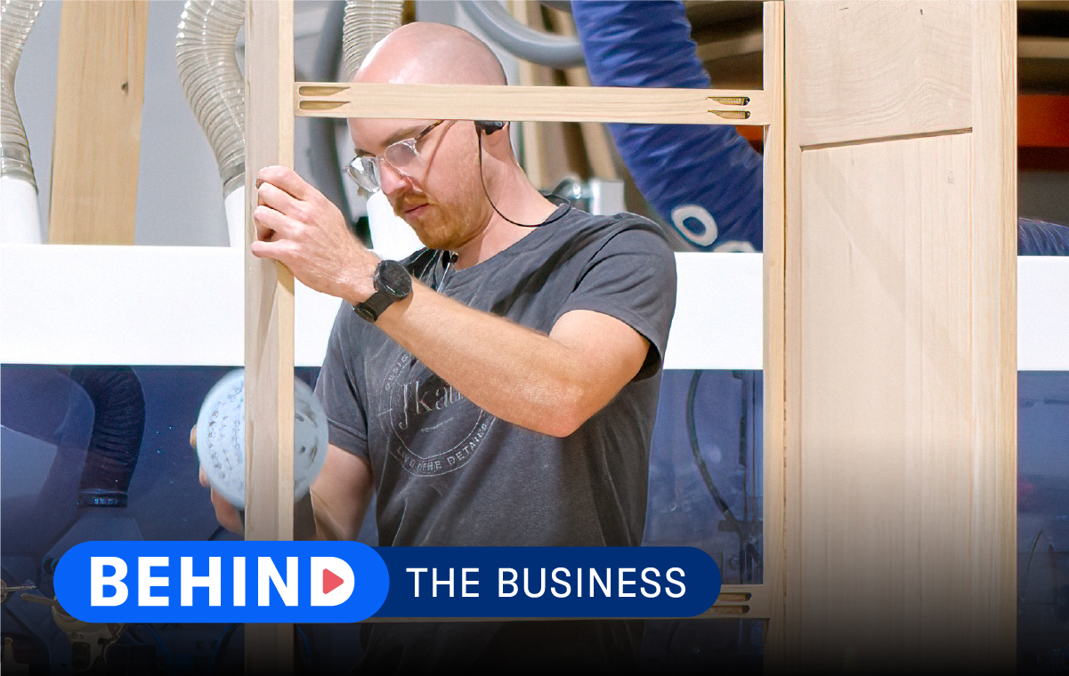 Construction worker building a cabinet in a shop with text that reads "Behind the Business."