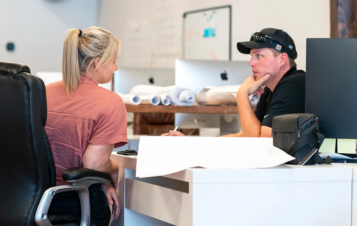 A man and a woman talking with blueprints on the desk in front of them