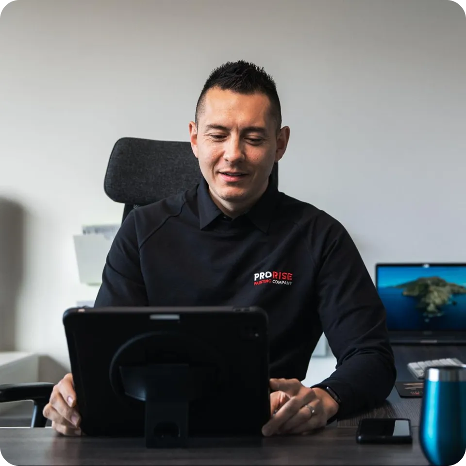 Man sitting at a desk working on a tablet, with a laptop and office supplies in the background