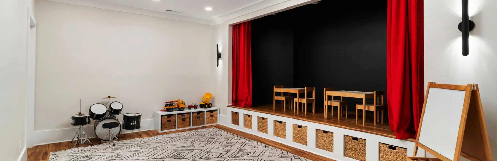 Children’s playroom with small stage, red curtains, toy drum set, and classroom-style tables