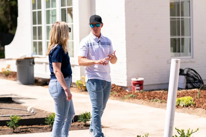 Two people discussing a construction project outside a white brick home with large windows and landscaping in progress.
