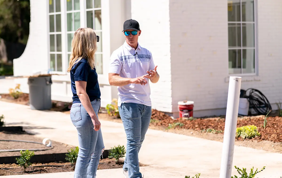 Two people discussing a construction project outside a white brick home with large windows and landscaping in progress.