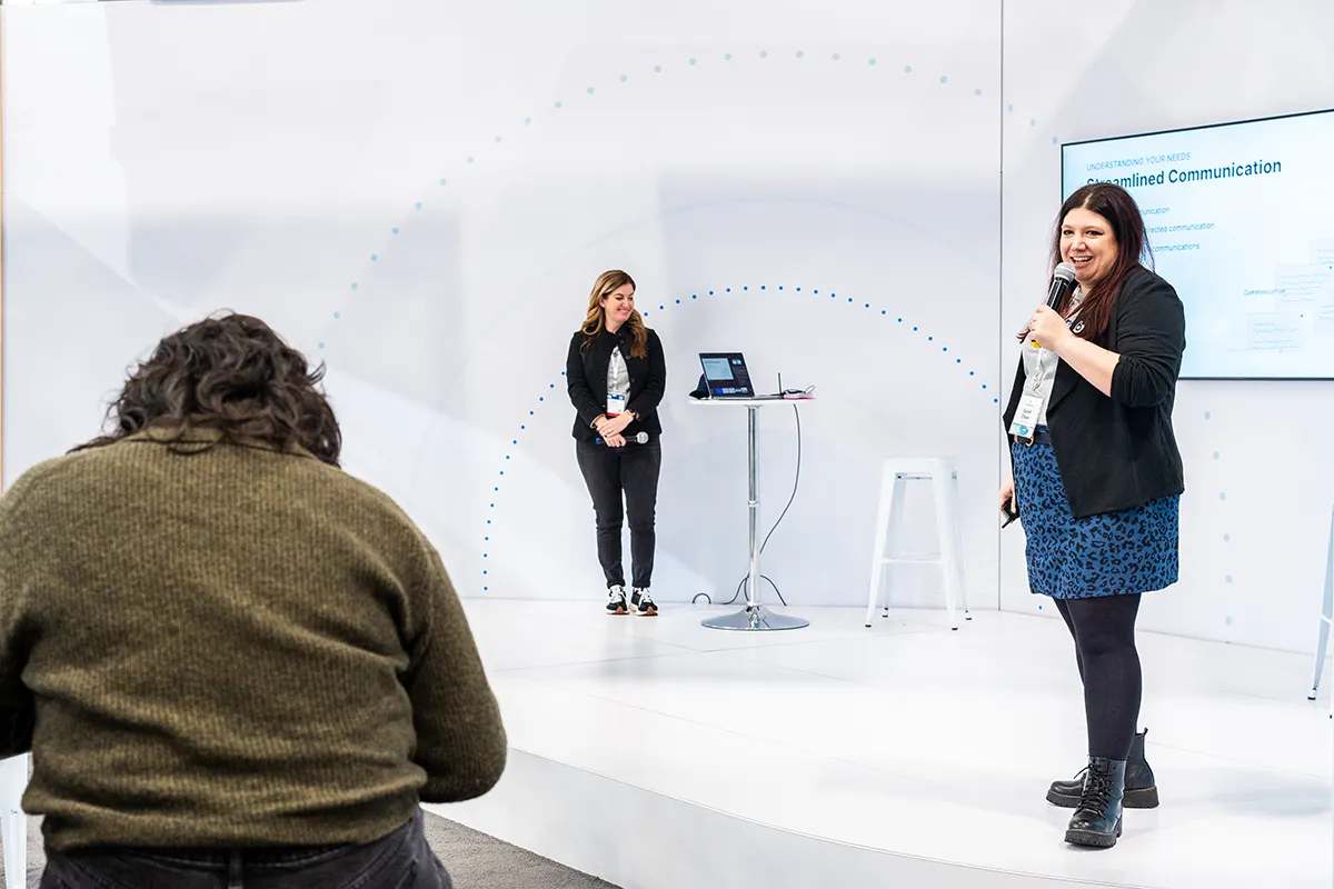 Presenter speaking on stage with microphone during a Buildertrend event, showing streamlined communication slide on screen.