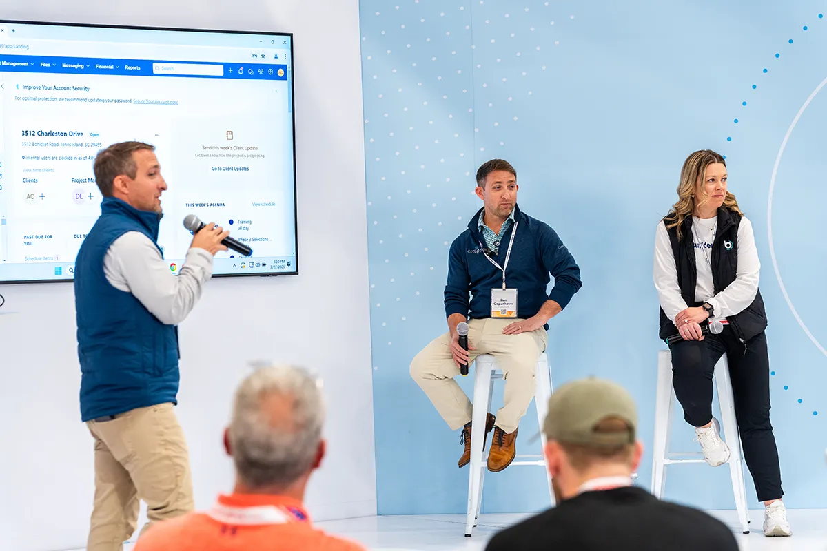 Speaker presenting on stage with microphone during a Buildertrend event, showing project management dashboard on large screen.