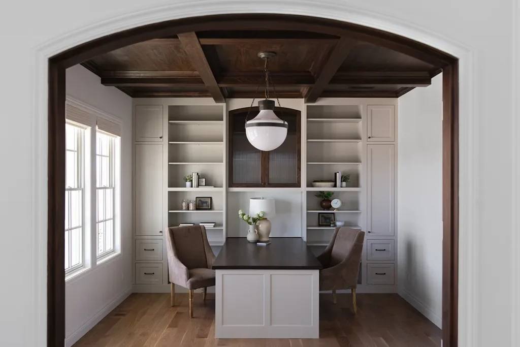 Home office with built‑in shelving, central desk, wood beam ceiling, and large windows for natural light.