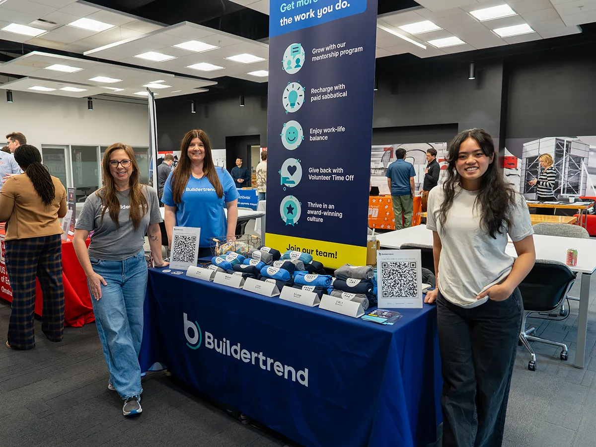Buildertrend booth at a career fair with a branded table display, promotional items, and representatives standing beside an informational banner.