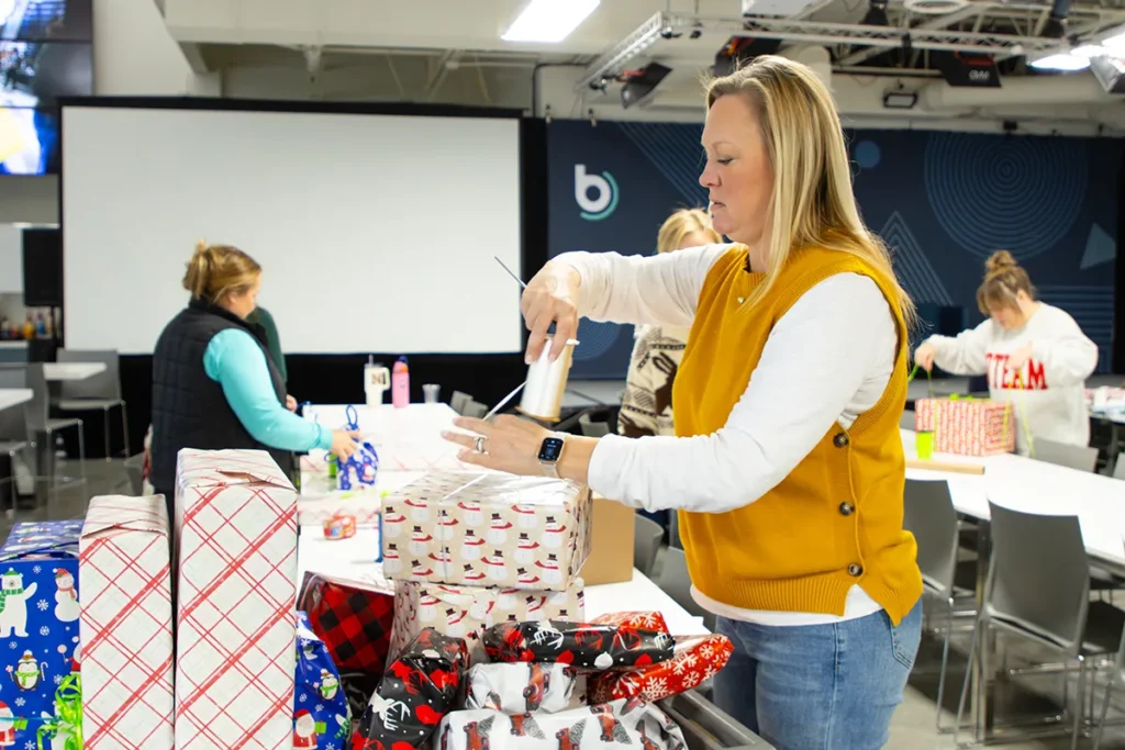 People wrapping gifts at tables during a volunteer event with boxes and holiday paper.