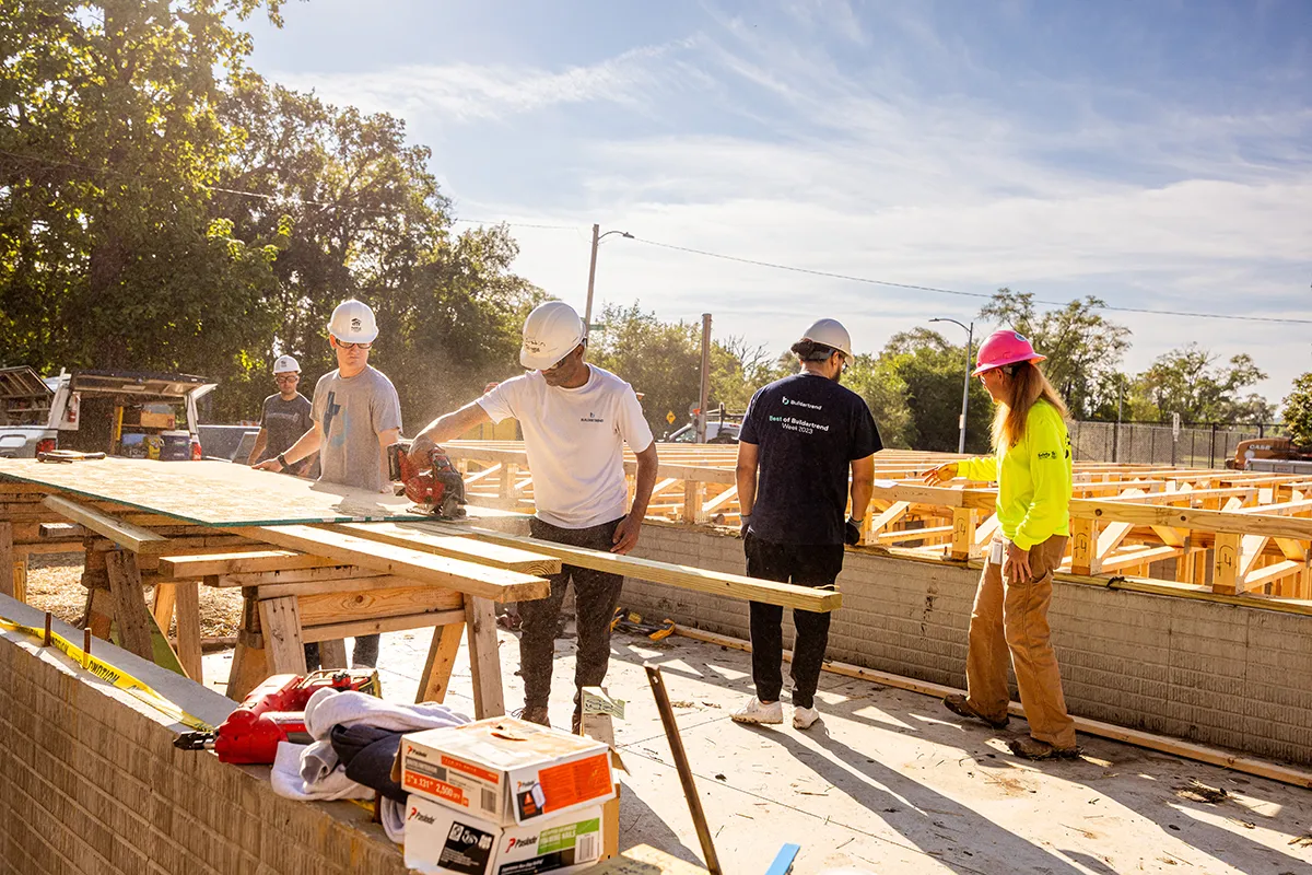 Construction crew working outdoors on a build site, cutting and measuring lumber while wearing hard hats and safety gear.