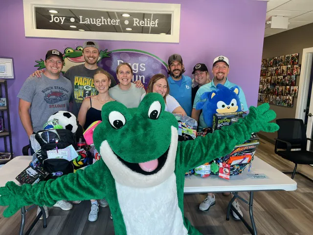 Team gathered around a table filled with donated toys and supplies, with a large frog mascot posing in front of the display.