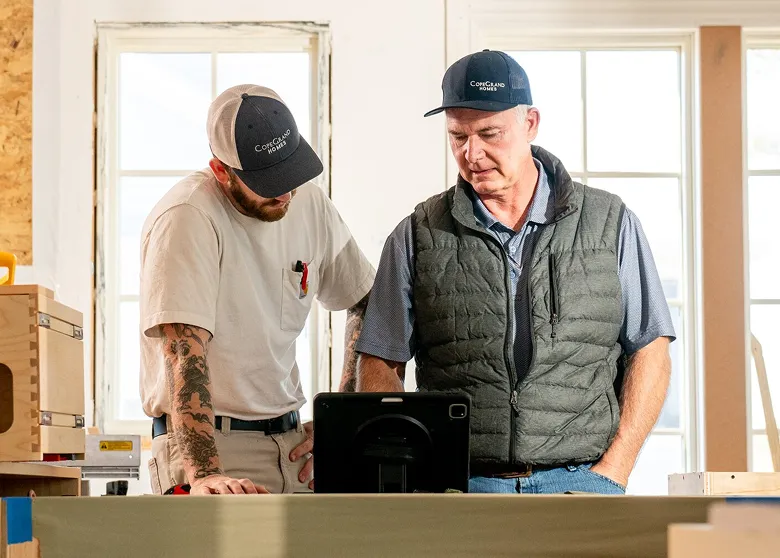 Two men wearing caps stand at a workbench in a construction or workshop setting, looking at a tablet device. The man on the left wears a light-colored shirt with a visible tattoo on his arm, while the man on the right wears a dark quilted vest over a blue shirt. Large windows and wooden materials are visible in the background.