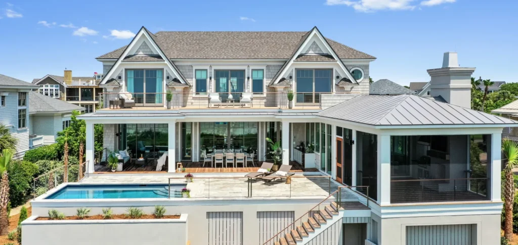 Modern two-story coastal home with cream and light blue exterior, featuring a swimming pool, covered patio areas, large windows, and manicured landscaping overlooking a residential neighborhood.