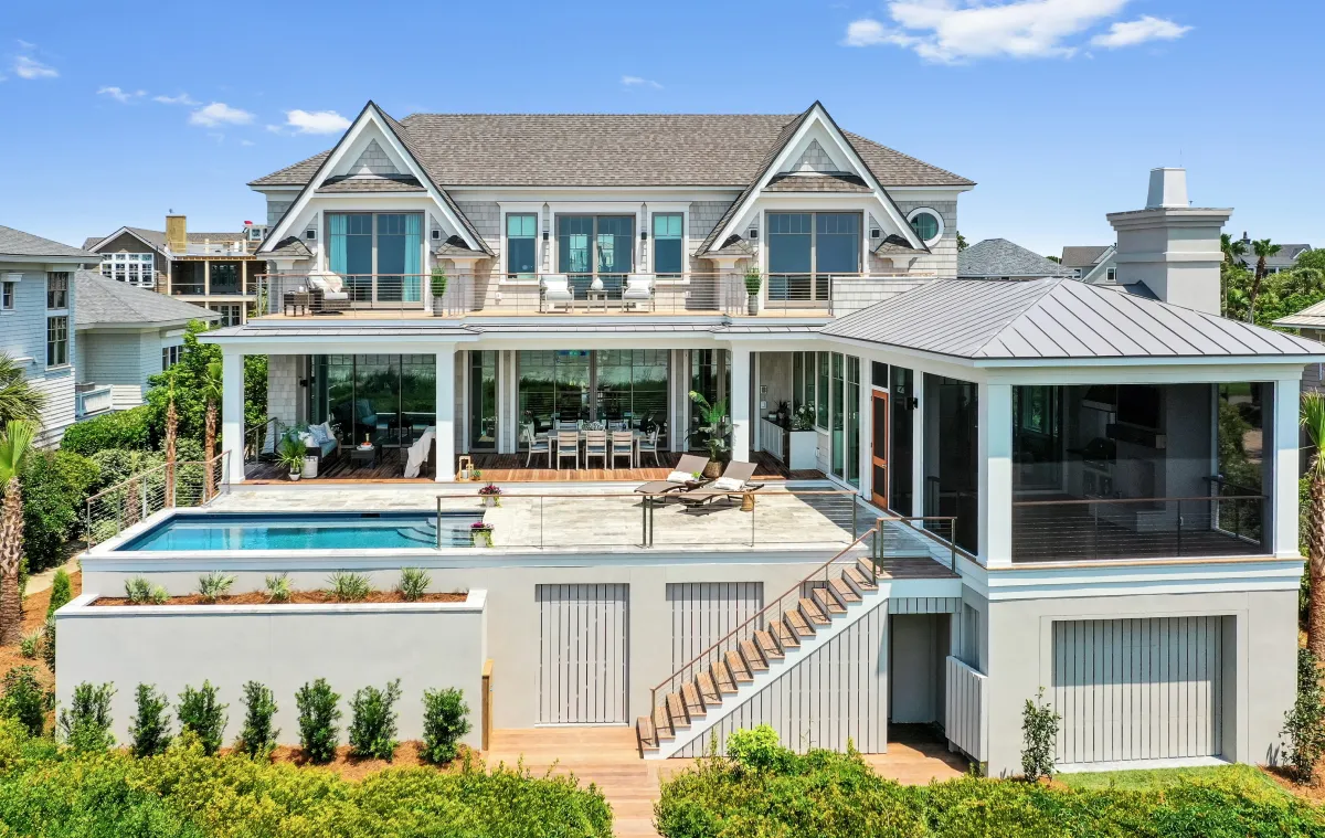 Modern two-story coastal home with cream and light blue exterior, featuring a swimming pool, covered patio areas, large windows, and manicured landscaping overlooking a residential neighborhood.
