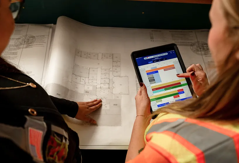 Two construction workers review blueprints and a project timeline on a tablet device at a job site table.