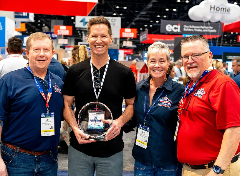 Four people posing together at a trade show or convention center, with the man in the center holding an award trophy while wearing a lanyard badge. Three others wearing lanyards and branded polo shirts stand beside him, smiling at the camera. Large displays and booths are visible in the background.