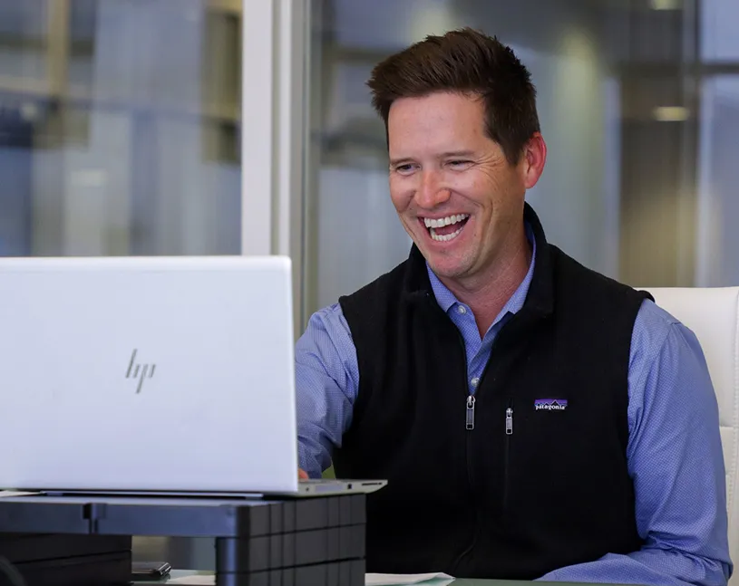 Dan Houghton smiles while working at a laptop in a modern office setting with large windows in the background.