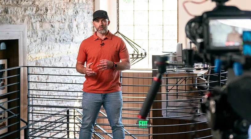 A man in a red polo shirt and black cap stands on an industrial metal platform, speaking to a camera during what appears to be a video recording or live stream in a brick-walled studio space.