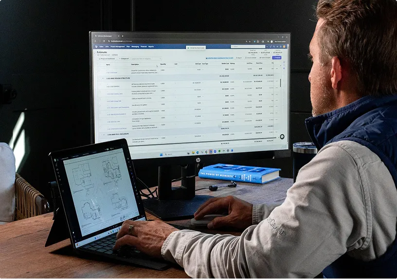 A man working at a desk views architectural floor plans on a tablet while monitoring a spreadsheet on a large desktop monitor, with a blue reference book nearby.