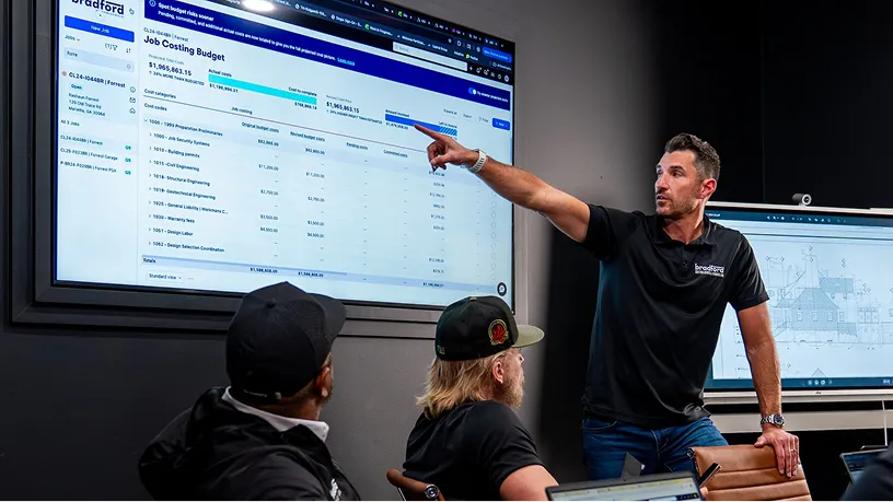 A man in a black shirt points to a large display screen showing a "Job Costing Budget" spreadsheet while two seated colleagues in black clothing observe the presentation.