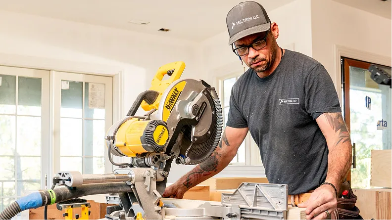 A man wearing a gray company t-shirt and cap operates a yellow and black miter saw in a bright indoor workshop setting, with large windows visible in the background.
