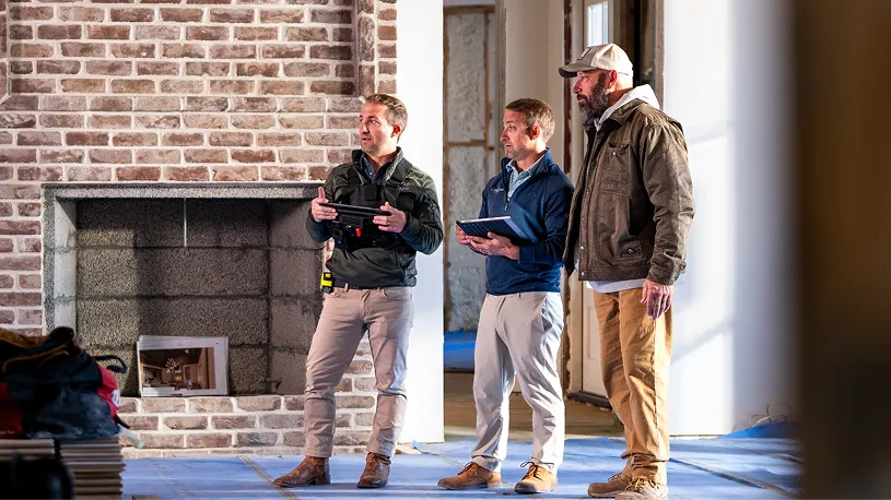 Three men discussing construction or renovation plans inside a brick-walled room with a fireplace, one holding a tablet device and another wearing a white hard hat.