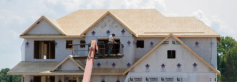 A two-story residential house under construction with white house wrap, exposed wooden framing, and workers on scaffolding installing roof shingles on a clear day.