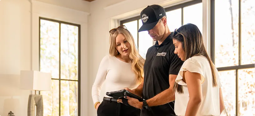 A uniformed service technician in a black cap and shirt shows a digital tablet to two women in a bright, modern home with large windows.