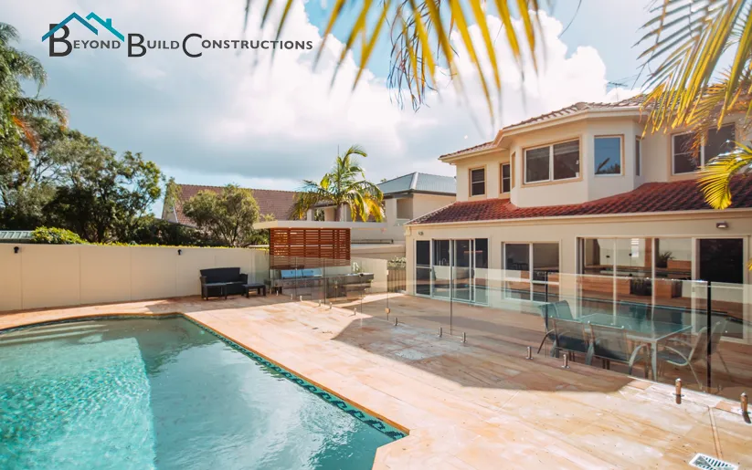 Luxury two-story residential home with swimming pool, modern glass fencing, wooden deck, and manicured backyard featuring palm trees and outdoor furniture, branded with Beyond Build Constructions logo.