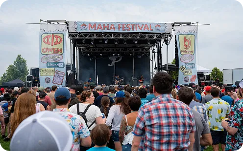 A large crowd of festival-goers gathered in front of a stage at the Omaha Festival, with sponsor banners visible on either side of the stage structure.