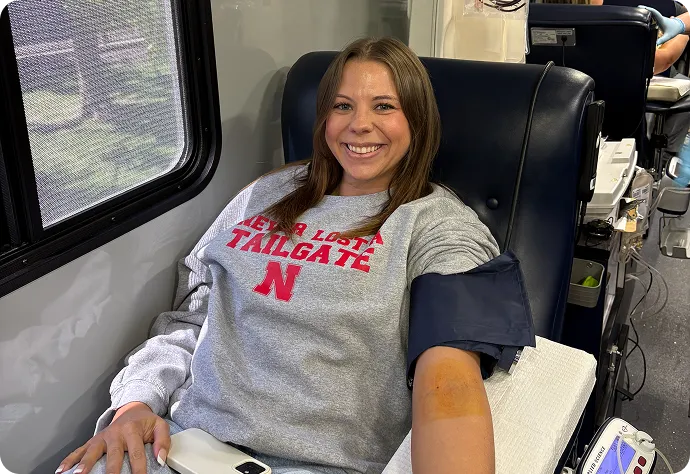 A smiling woman wearing a gray sweatshirt with red text sits in a black chair inside what appears to be a vehicle or bus. She has a blood pressure cuff on her arm and is looking at the camera.