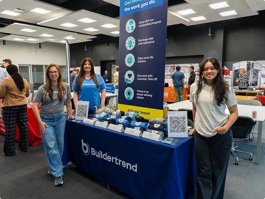 Buildertrend recruitment booth at a job fair with team members standing behind a blue table displaying promotional materials and branded merchandise, featuring a banner highlighting company benefits including mentorship, paid sabbatical, work-life balance, and volunteer time off.