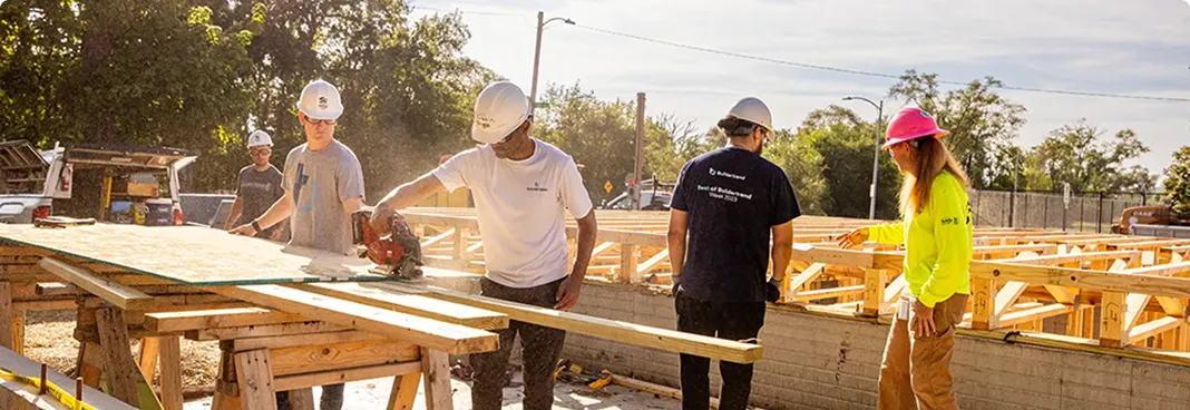 A construction crew of five workers wearing hard hats and safety gear work together at an outdoor building site, examining wooden framing and materials on a work table with residential construction in progress in the background.