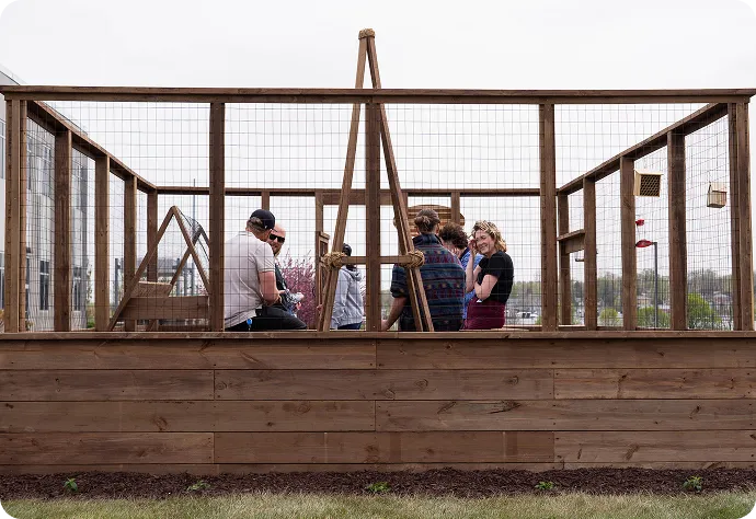 A group of people inside a large wooden greenhouse with mesh panels, gathered around gardening equipment during what appears to be a community gardening activity or workshop.