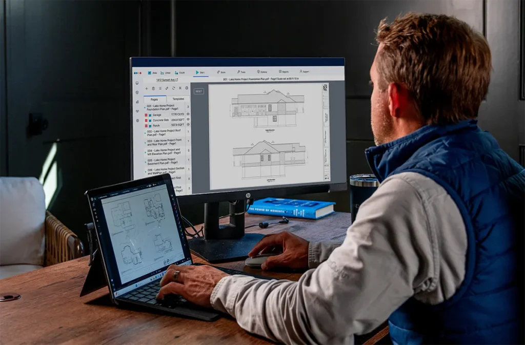 Buildertrend Takeoff Paid Campaign LP 6 A man wearing a blue vest works at a desk with a tablet and monitor displaying architectural house plans and elevation drawings.