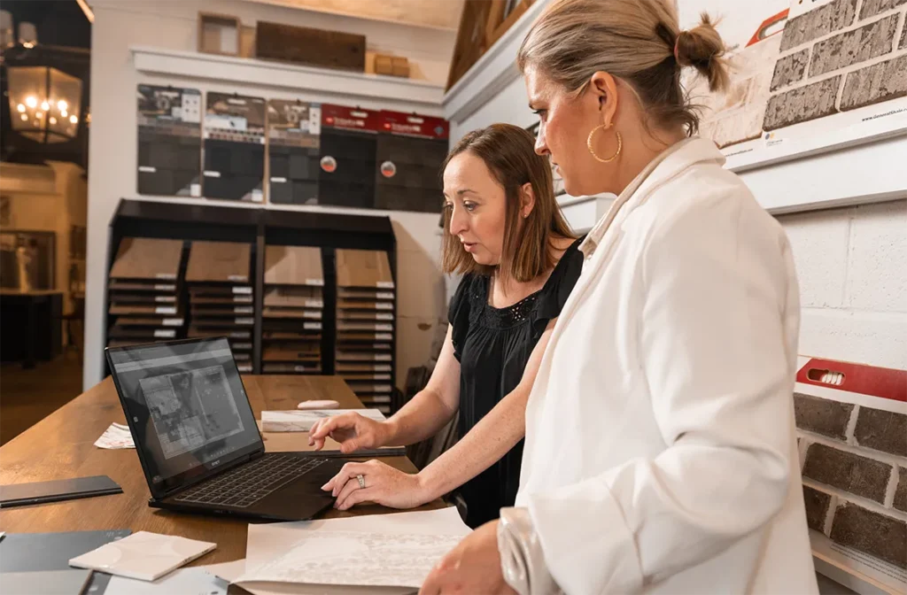 Buildertrend Takeoff Paid Campaign LP 7 Two women examining a laptop together in a flooring showroom with tile and wood samples displayed on the walls and in storage units behind them.