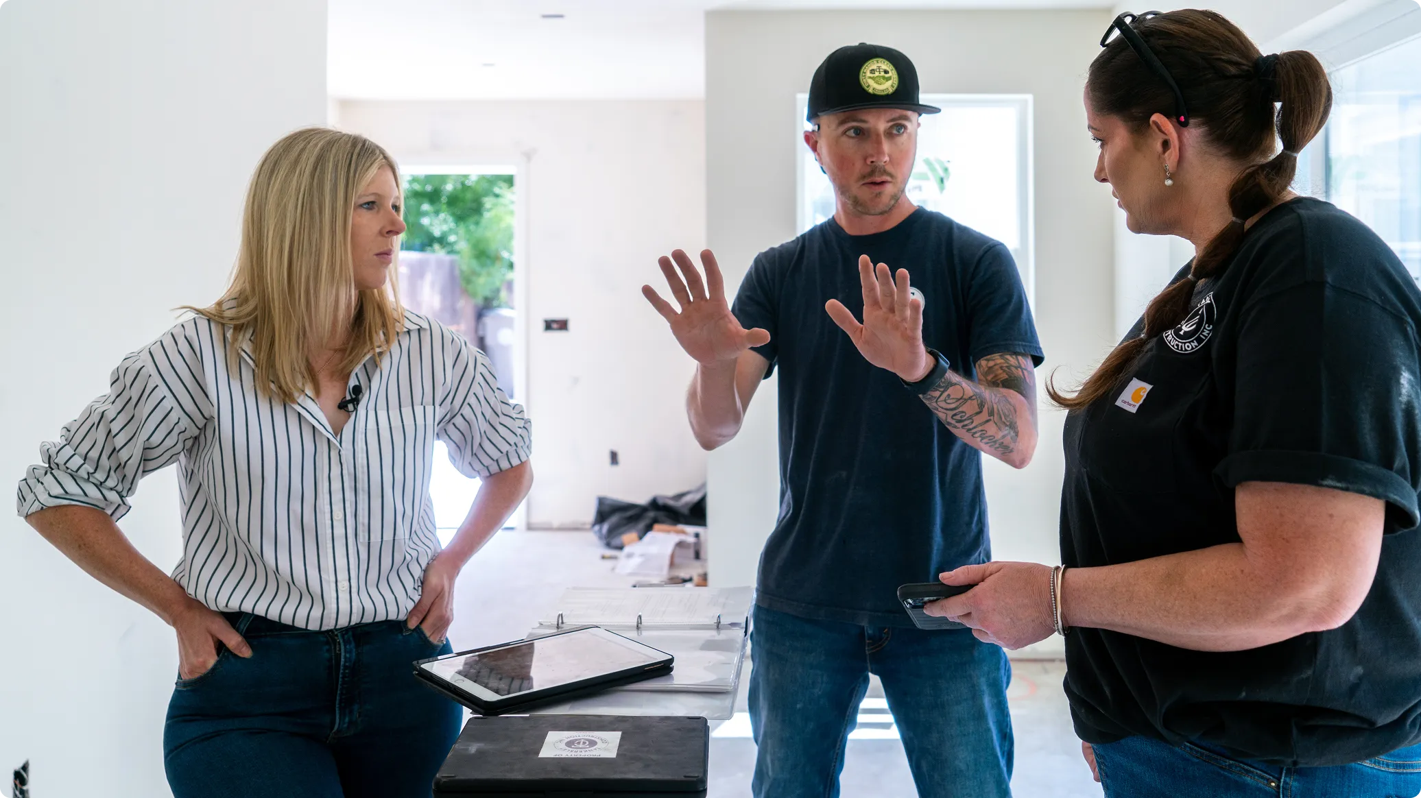Three people having a discussion in a bright, modern interior space. A woman in a pinstriped shirt stands on the left with a tablet, while a man in a navy cap and dark shirt gestures while speaking in the center, and another woman in a dark shirt with a logo holds a phone on the right.
