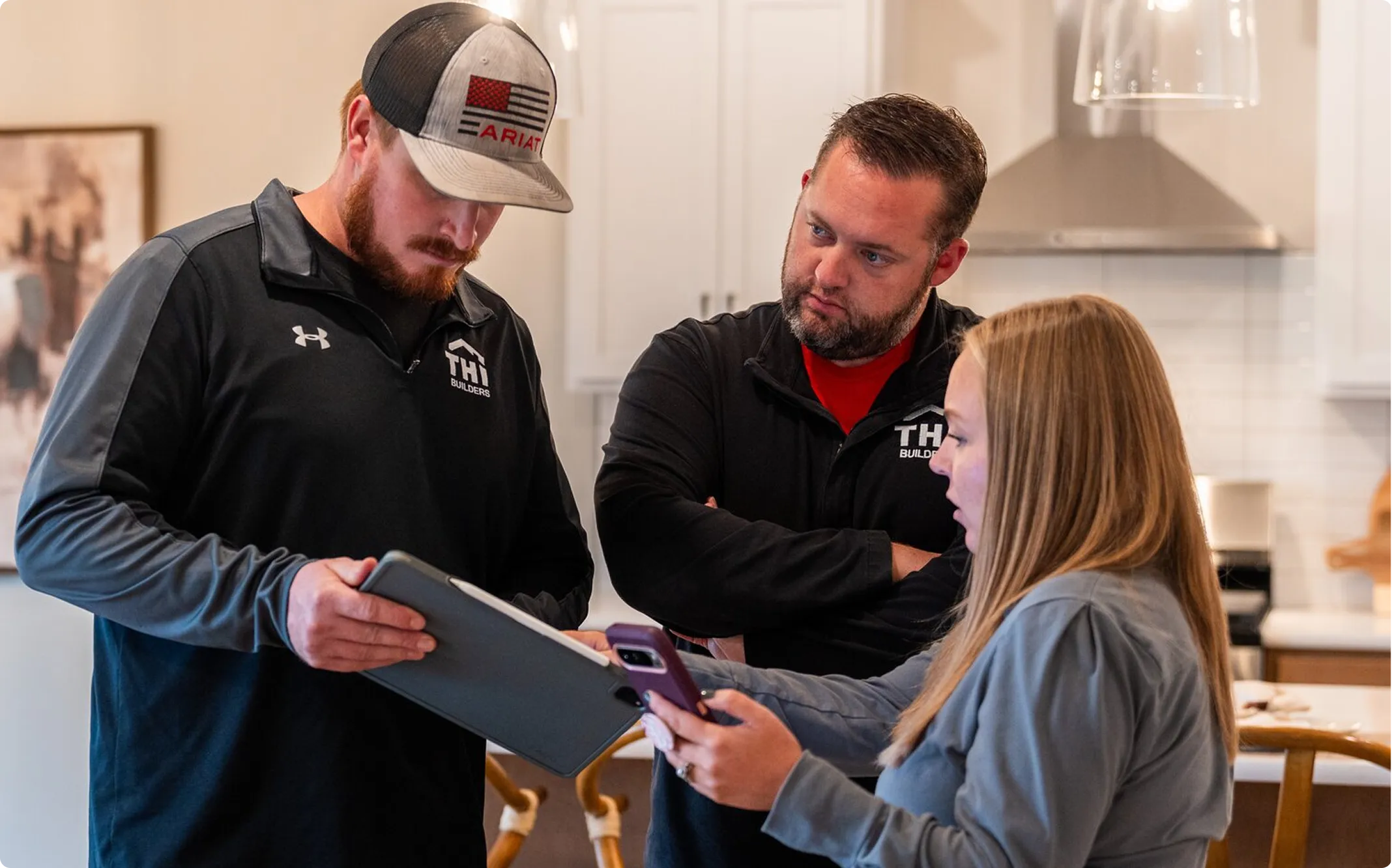 Three people reviewing construction or renovation plans on a tablet in a kitchen interior, with two men wearing THI Builders branded clothing and a woman holding a mobile device.