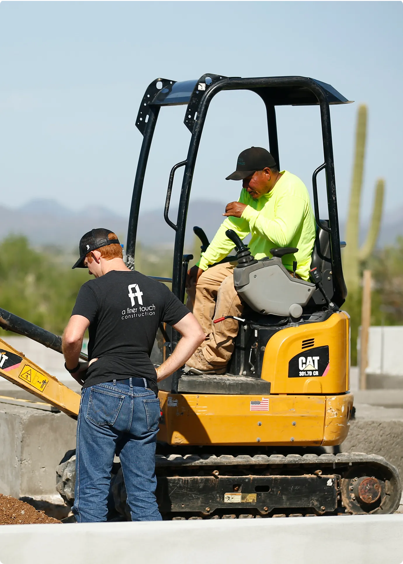 Two construction workers operating a yellow CAT mini excavator on a job site, with one worker in the cab and another standing beside the machine, desert landscape with saguaro cacti visible in the background.