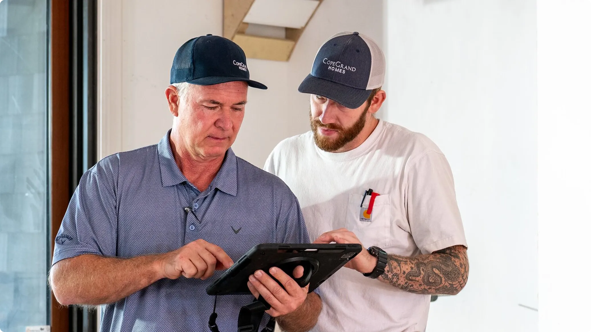 Two men in casual work attire examining a tablet device indoors. The man on the left wears a blue polo shirt and blue cap, while the man on the right wears a white t-shirt with a logo and a dark cap. Both are focused on the tablet screen in a bright, modern interior space.