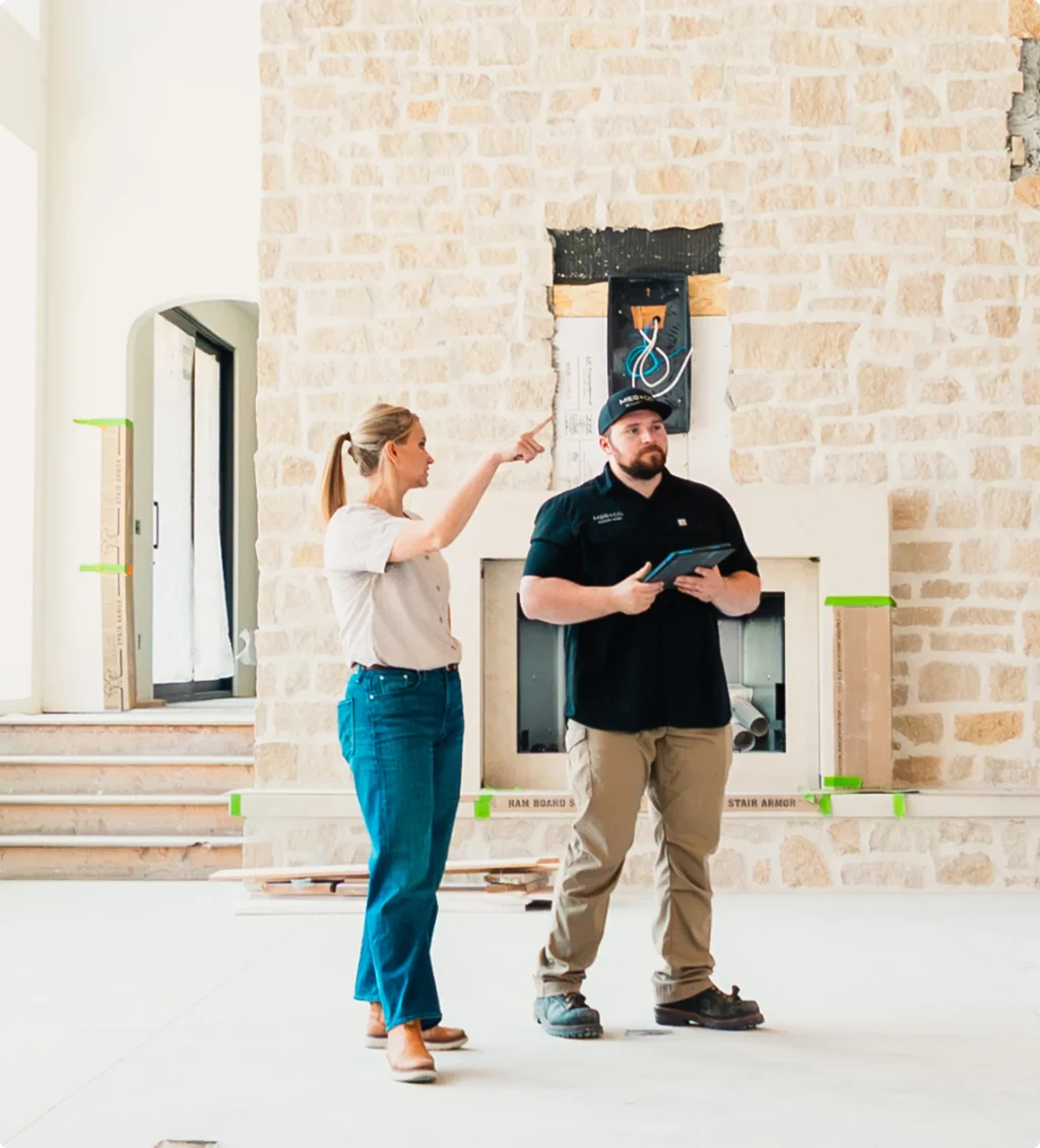 A woman in a white shirt and blue jeans points toward electrical wiring in a brick wall while a man in a dark polo shirt holding a tablet stands beside her during what appears to be a home construction or inspection walkthrough.