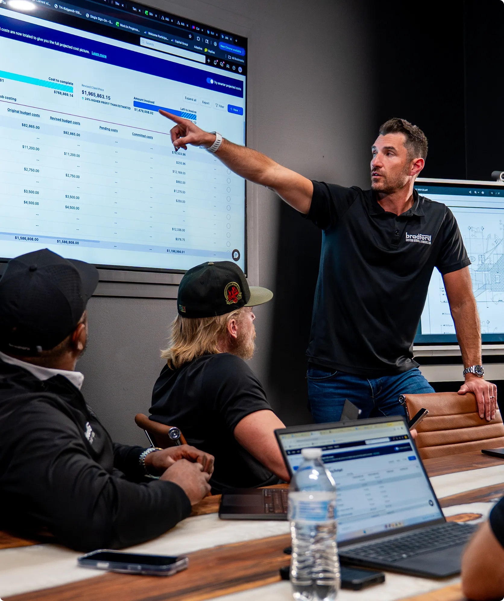 A man in a black Bradford Building polo shirt presents financial data on a large screen to two seated colleagues during a business meeting, pointing to a cost breakdown chart while a laptop displays related information on the wooden table.