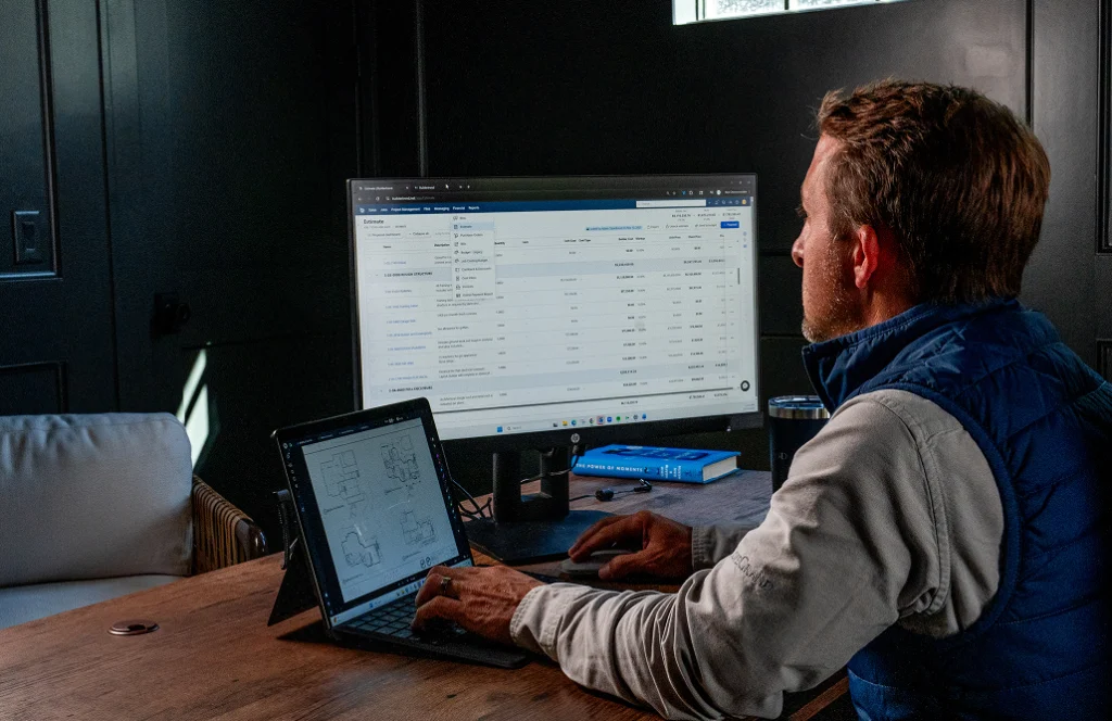 Contractor reviewing a project estimate on a desktop monitor while a tablet showing floor plans sits nearby on his desk