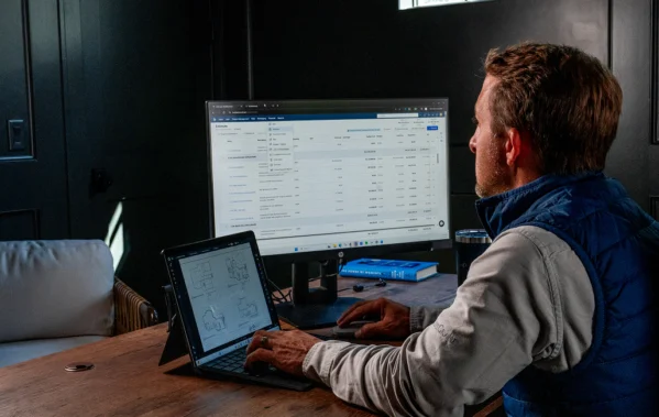 Contractor reviewing a project estimate on a desktop monitor while a tablet showing floor plans sits nearby on his desk