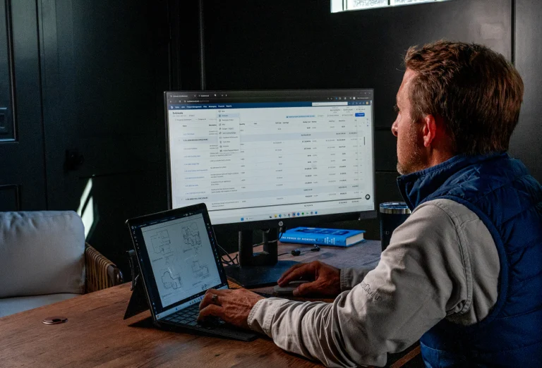 Contractor reviewing a project estimate on a desktop monitor while a tablet showing floor plans sits nearby on his desk