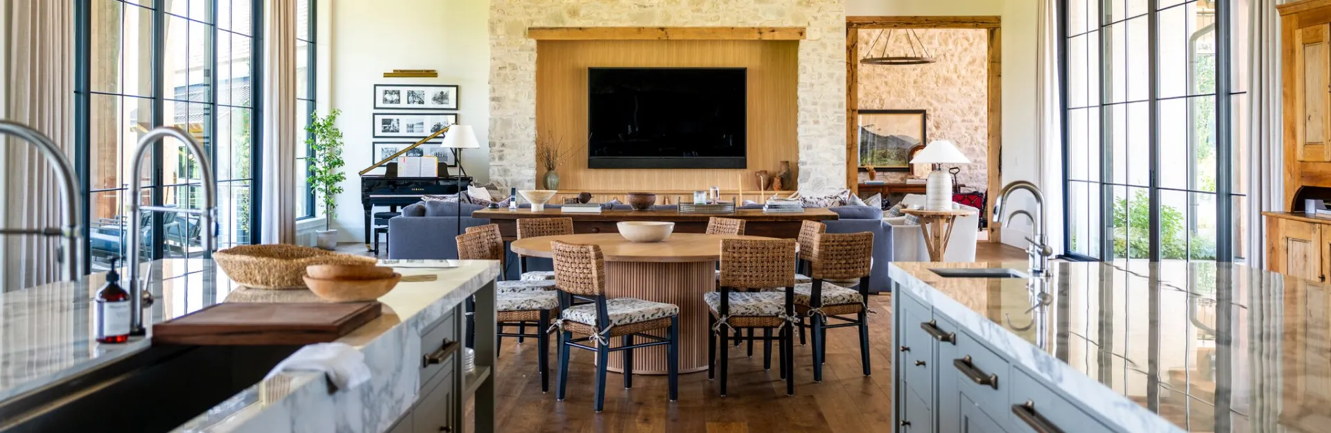 Scaling to a $50 Million Luxury Builder 2 Modern open-concept kitchen and living room with marble countertops, wooden dining table with woven chairs, large windows with natural light, stone fireplace with TV, and contemporary furnishings throughout.