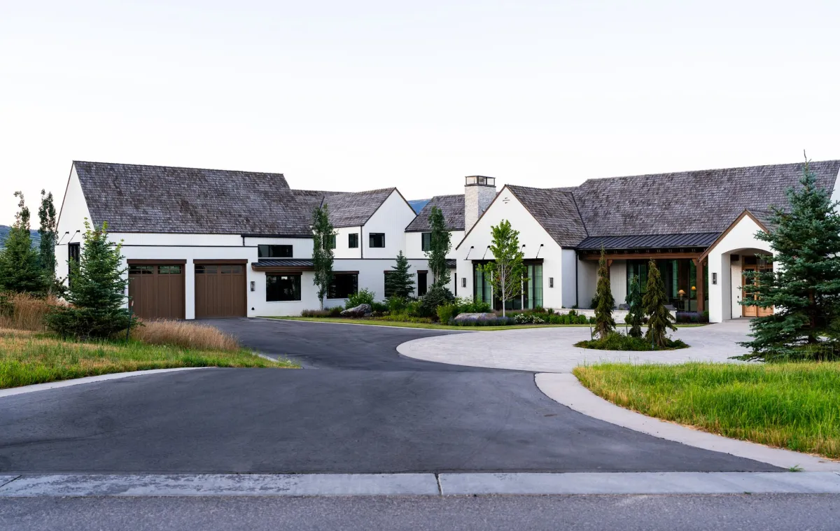 Scaling to a $50 Million Luxury Builder 1 A modern white farmhouse with dark gray roofing and brown garage doors, featuring multiple gables and a central circular driveway with landscaping, surrounded by green lawn and evergreen trees.