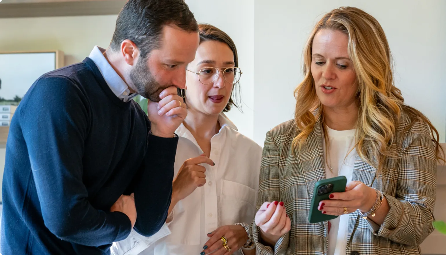 Three professionals in an office setting reviewing content on a mobile device, with a man in a dark sweater and blue shirt on the left, a woman in glasses and beige top in the center, and a woman in a tan plaid blazer on the right holding the phone.